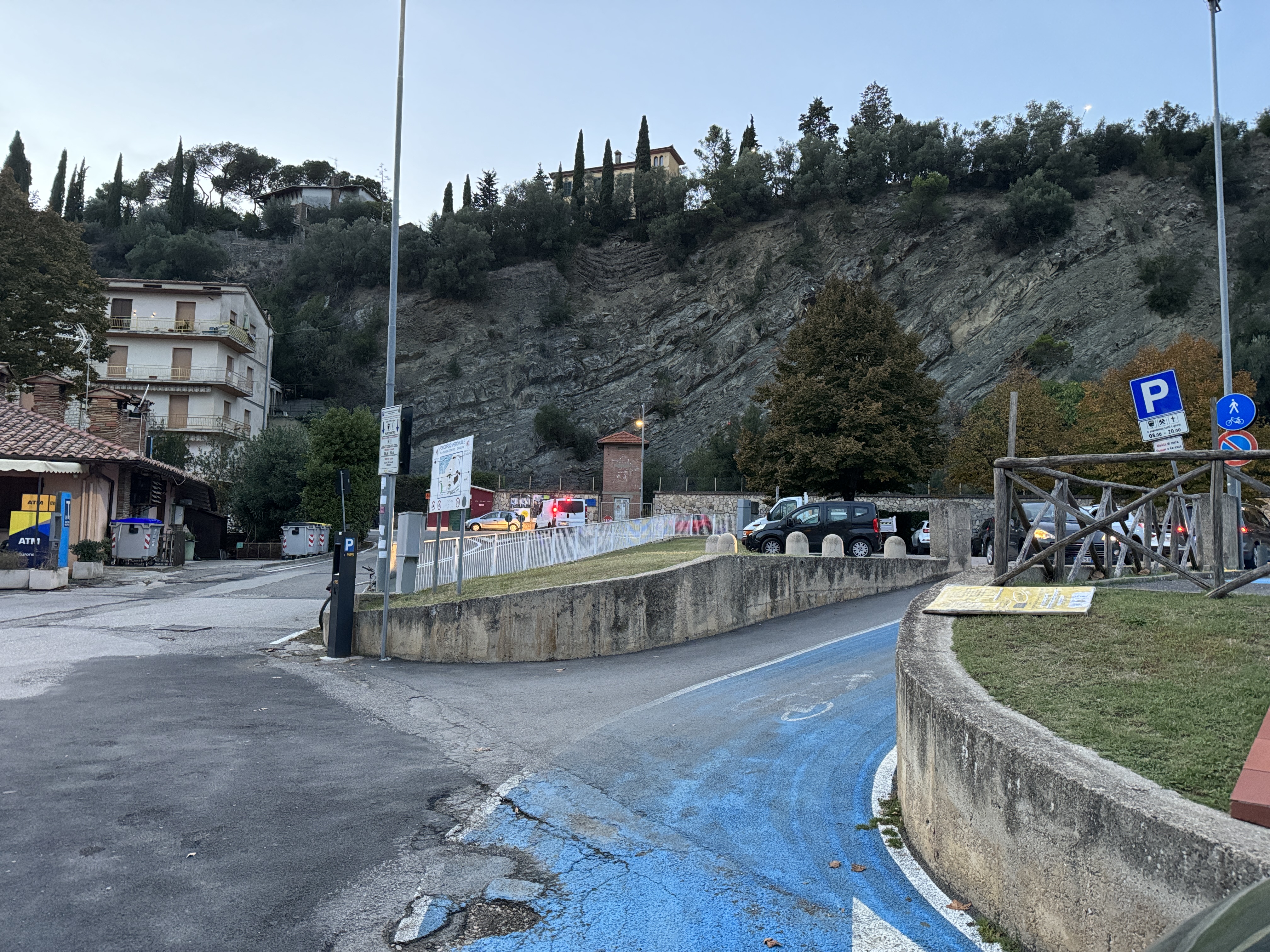 Blauer gepflasterter Radweg aus dem Parkplatz heraus. Biegt nach rechts ab. Auf der linken Seite: Gebäude und Fußgängerwegschilder